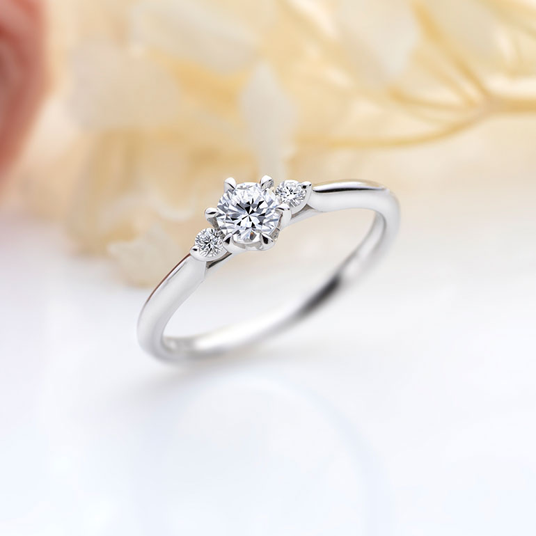 センターのダイヤモンドに花を添えるようにセットされた両サイドのメレダイヤモンドがより華やかな印象にしてくれます。正統派なデザインですが、サイドから見えるダイヤモンドを留めているシャトンデザインが繊細で可愛らしさも演出します。